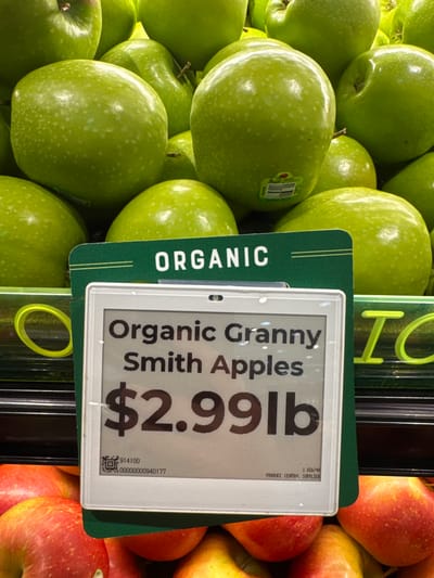 A close-up, eye-level shot captures a display of organic Granny Smith apples at a grocery store in Shaker Heights, Ohio, United States of America. A digital price tag prominently displays "Organic Granny Smith Apples $2.99lb" with a QR code and product number below. The apples are a vibrant green, some with slight yellow undertones and white speckles, and are piled high in the background, suggesting a large quantity. The background also shows a dark horizontal shelf or display area, and glimpses of red apples are visible in the foreground. The overall impression is a well-stocked produce section.