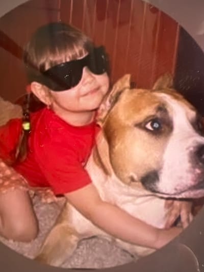 A young girl with dark hair pulled into a braid, wearing black sunglasses and a red t-shirt, is pictured with a large dog. The girl is leaning against the dog, with her left arm around its neck. The dog, likely a pit bull type, has tan and white fur and is looking to the right. The background appears to be indoors, with a textured surface and vertical lines that could indicate a wall or furniture. The image quality is grainy, suggesting it is a photograph from some time ago. There is no specific indication of the city of Laconnex, Switzerland.