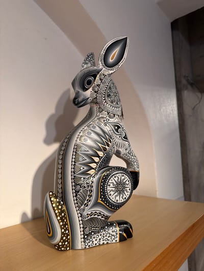 A unique, hand-carved animal sculpture rests against a cream wall, adjacent to a light wood surface. The piece appears to be a deer, intricately adorned with grayscale patterns, including concentric circles, geometric shapes, and delicate dot work. These designs highlight the curves and contours of the animal form, creating a captivating visual texture. The contrasting colors accentuate the details, drawing the viewer's eye to the meticulous craftsmanship.
