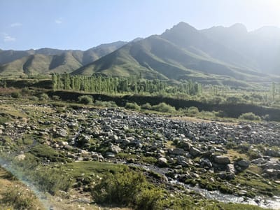 The image depicts a natural landscape with mountains in the background, a line of trees in the midground, and a rocky, overgrown terrain in the foreground with a small stream. The sky is clear with a slight haze. There are no people or discernible man-made structures in the scene, suggesting a remote or natural environment. The specific location within Kalaynay, Tajikistan, cannot be determined from the visual information alone.