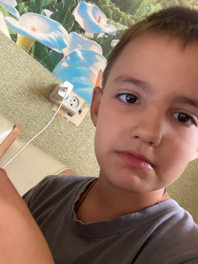 In a close-up, selfie-style shot, a young boy with brown hair and brown eyes is pictured. He is wearing a gray t-shirt. His left arm is visible in the foreground, partially obscuring the view. In the background, a wall features a floral wallpaper with white calla lilies and green leaves. To the left of the boy, a wall socket with a white plug inserted into it is visible against a textured, olive-green surface. The setting appears to be indoors, possibly a residential room in Krasnovka, Russia, given the context provided.