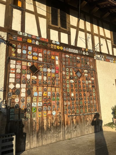 This image captures the exterior of a building in Rafz, Switzerland, illuminated by strong, low-angle sunlight. The primary feature is a large, weathered wooden double gate, extensively adorned with hundreds of small, multicolored plaques or medals of various shapes, including oval, rectangular, circular, and shield-like forms. Many of these plaques display equestrian motifs such as horses and riders. Two small, dark-framed, diamond-shaped windows are set into the upper sections of the gate panels. Above the gate, the building's facade exhibits traditional half-timbered construction with white infill panels and dark wooden beams. A window with dark louvered shutters is visible on the upper left. The ground in front of the gate is paved. A distinct human silhouette, appearing to hold a camera or phone, is cast onto the lower right gate panel and the ground, indicating the presence of a photographer. To the far left, a light-colored plastic crate containing bottles is partially visible in shadow. A small potted plant is positioned on the ground to the far right. No other individuals or direct interactions are visible.