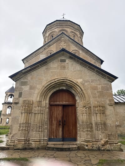 The image shows the exterior of the Nikortsminda Cathedral in Georgia. The cathedral is constructed from light-colored stone blocks and features intricate carvings around the arched doorway and along the roofline. A large wooden door is visible within the arch. To the left, a section of the octagonal drum of the main dome is visible, also adorned with decorative carvings. In the background, a smaller bell tower with a dark roof and arched openings can be seen. The sky is overcast, suggesting a cloudy or rainy day, and the ground is covered with stone paving. Green grass is visible to the right and in the lower background. The architectural style appears to be medieval Georgian. There is no visible text in the image.