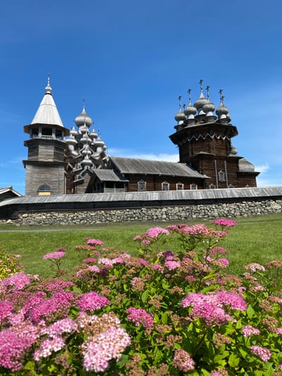 The image captures a bright, sunny day with a clear blue sky over the Kizhi Pogost in Boyarshchina, Russia. In the foreground, a lush green lawn is adorned with clusters of pink and white flowers, with some in full bloom and others in bud. Behind the flowers, a low stone wall encloses a complex of wooden churches. The larger of the two prominent churches, the Church of the Transfiguration, features multiple bulbous, silver domes, each topped with a cross. Adjacent to it stands the bell tower, also constructed of wood and topped with a conical spire. The churches exhibit intricate wooden architecture, typical of traditional Russian style. The entire complex is surrounded by a wooden fence. The scene is serene, highlighting the historical and architectural significance of the location under a vibrant sky.