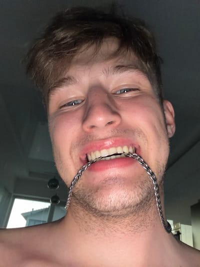 A close-up, slightly upward-angled selfie shows a young man with light brown, tousled hair, blue eyes, and a light stubble. He is biting down on a thick, silver-colored metal chain, holding it between his teeth. His lips are slightly parted, revealing white teeth. The background is dark and out of focus, with some blurry shapes and lighting that suggest an indoor setting. There are reflections visible in the background that appear to be of windows with a building structure outside, and possibly some hanging lights. The overall impression is of a casual, self-taken photograph. The image does not contain any elements that can be used to determine a location in Vanino, Russia.