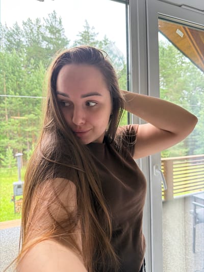 The image shows a young woman lying down indoors, with her arm raised above her head. She is wearing a brown long-sleeved shirt. Her hair is long and brown, falling around her face and shoulders. She is looking towards the camera with a relaxed and playful expression. The background appears to be a window overlooking a lush green outdoor setting, likely a yard or garden with trees. The lighting suggests it is daytime, possibly overcast due to the soft light. The scene conveys a sense of casual comfort and personal moment captured.