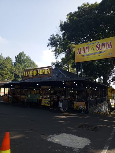 This image captures a vibrant roadside restaurant complex, identified by prominent signage as "WARUNG NASI ALAM SUNDA," which translates to "Alam Sunda Rice Stall." The language used on the signs is Indonesian, with "Untuk Semua Kalangan" meaning "For All People," and references to "SATE KAMBING" (Goat Satay) and "IKAN & ALAM BAKAR" (Grilled Fish & Chicken/Nature). This linguistic and culinary context strongly indicates the establishment is located in Indonesia, directly contradicting the provided location of Weddagala, Sri Lanka.
The scene is presented under a bright, clear sky, with numerous lush green trees forming a dense background and surrounding the building, suggesting a natural or semi-rural setting. The main structure is a single-story building with a dark grey, ridged roof, featuring an open-air or semi-open design. Beneath this roof, multiple food stalls or counters are arranged, illuminated by numerous small, exposed light bulbs hanging from the ceiling structure.
In the foreground, an asphalt road appears dark grey with some lighter patches, possibly from water or reflections. A white line is partially visible on the bottom right, and a rectangular metal drain grate is embedded in the road in the lower right quadrant. An orange and yellow traffic cone is partially visible in the bottom left corner.
The restaurant itself is elevated from the road level, with a stone-faced retaining wall visible on the lower right, marked with distinctive yellow and black hazard stripes. A dark-colored metal railing runs along the edge of the elevated platform. Within the stalls, various items are visible, including food display units, what appears to be fresh produce (like durians), cooking equipment, and promotional banners for specific dishes. Several individuals are present inside the restaurant area, with at least one person in a dark uniform visible on the far left, standing near a stall. Other figures are less distinct, engaged in general activity typical of a food service environment. A large yellow billboard sign on tall poles prominently displays the "ALAM SUNDA" branding on the right side of the image, while a smaller, similarly branded sign sits atop the restaurant's roof. A diamond-shaped road sign, also advertising "ALAM SUNDA," is visible on the far right near the retaining wall.