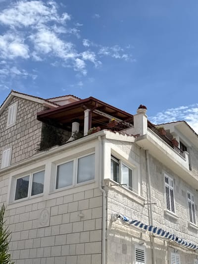 The image shows a white stone building with a red tiled roof and a wooden pergola. The building is located on the Dalmatian coast of Croatia, in the town of Dugi Rat. The building is two stories tall and has a balcony on the second floor. The windows are all closed and have white frames. There is a blue and white awning over the ground floor windows. There are plants in pots on the balcony. The sky is blue and there are some white clouds. The building is located at coordinates 43.446, 16.636.