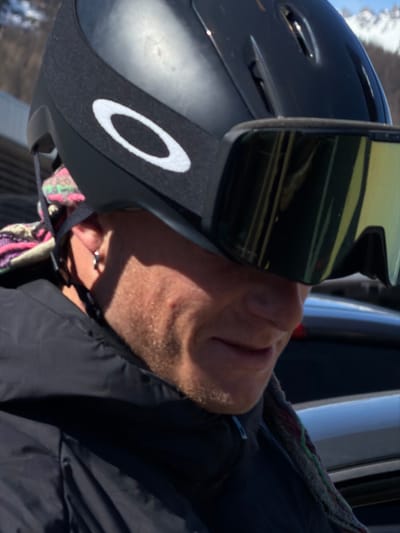 The image captures a close-up, angled view of an individual in La Salle-les-Alpes, France, positioned outdoors under direct sunlight. The subject, appearing to be an adult male, wears a black ski helmet featuring a white Oakley logo on its left side and large, reflective yellowish-green ski goggles that obscure the eyes. Stubble is visible on the subject's cheek and jawline, and a small silver hoop earring is present in the visible left ear. A multi-colored patterned fabric, displaying shades of pink, green, and purple, is visible around the neck area, beneath the helmet strap. The individual is dressed in a black, possibly technical, jacket with a visible light blue zipper pull at the collar. The background is out of focus, revealing the tops of dark-colored vehicles behind the subject and snow-covered mountain peaks in the upper right.