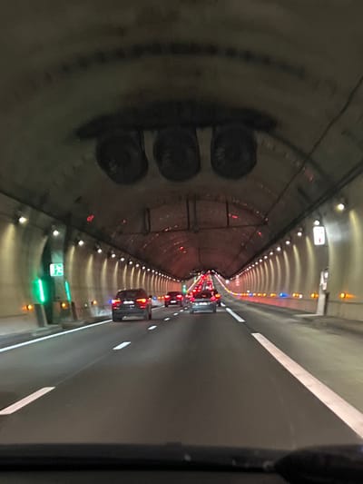 A slightly tilted perspective down a tunnel reveals a line of cars with illuminated tail lights. The tunnel's curved walls are lined with evenly spaced lights, diminishing into the distance. On the left, a dark, cylindrical shape dominates the foreground, possibly an element of the tunnel's infrastructure. The right side exhibits a smooth, light-colored surface, likely a walkway or wall. A faint green sign is visible in the lower center of the tunnel.