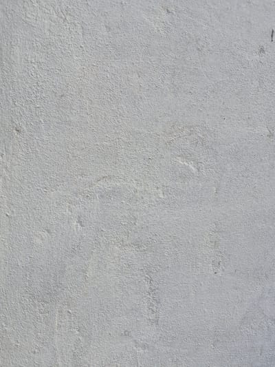 This is a close-up, overhead shot of a lightly textured, pale gray surface. The texture appears granular, suggesting it could be concrete, plaster, or a similar material. There are subtle variations in shade and some faint, irregular marks or smudges across the surface. In the top left corner, a small, dark, elongated object or shadow is visible. The lighting suggests it is daytime, and the overall impression is of a mundane, outdoor surface, possibly a wall or pavement. No people, objects, or text are clearly discernible. The context provided indicates the location is in Қанабек, Kazakhstan.