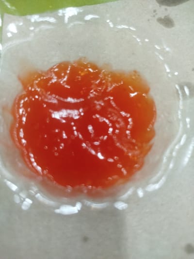 An overhead close-up captures a roughly circular mass of bright orange-red viscous substance, exhibiting a glossy surface with numerous small white reflections, consistent with a condiment or jam. This substance is contained within a translucent, thin, flexible material, likely a small plastic film or cup liner, which forms a distinct scalloped edge around its perimeter. The container rests on a flat, light-beige or off-white textured surface. A blurred green element is visible in the upper-left quadrant of the frame. The scene is static, with no human subjects or active interactions present. The location is Matangjareung, Indonesia.