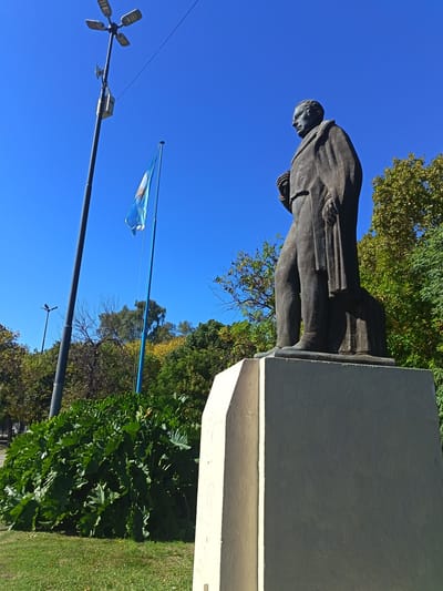The image presents a low-angle view of a bronze statue of a man situated in an outdoor public space under a clear, brilliant blue sky. The statue depicts a male figure in a formal coat, standing with his left hand holding what appears to be a book or scroll. It rests on a tall, light-colored, angled stone pedestal, partially visible in the lower right.
To the left of the statue, a tall, dark street light pole ascends towards the sky, topped with a modern fixture featuring four rectangular luminaires. A small, light-colored box is attached to the pole below the lights. Further to the left, a blue and white Argentinian flag, with its prominent "Sun of May" emblem, flies from a flagpole, caught gently by the breeze.
The mid-ground is filled with abundant green vegetation. Large, lush green bushes with broad leaves are prominent in front and to the left of the statue, while a dense canopy of trees with varying shades of green and some yellowish foliage forms the background. The foreground consists of well-maintained green grass.
The scene is bathed in bright sunlight, indicated by the crisp shadows and vivid colors, suggesting a clear day, likely in the morning or early afternoon. The overall setting is that of a park or public square in Remedios de Escalada, Argentina, marked by the presence of the national flag and civic monument. No specific activity is occurring, and no text is visible on the statue or its pedestal.