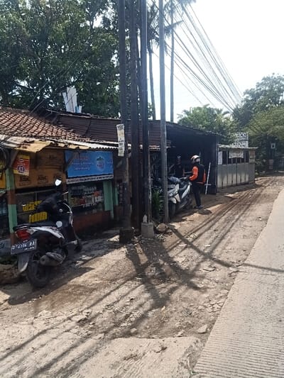 A weathered roadside scene features a couple of parked motorbikes near a small shop with a worn, corrugated metal roof. Utility poles stand nearby, casting long shadows across the uneven, dirt-patched road surface. In the distance, another shop or business establishment is visible, while trees and power lines dominate the background. A person in a helmet and an orange jacket is standing near the motorbike.