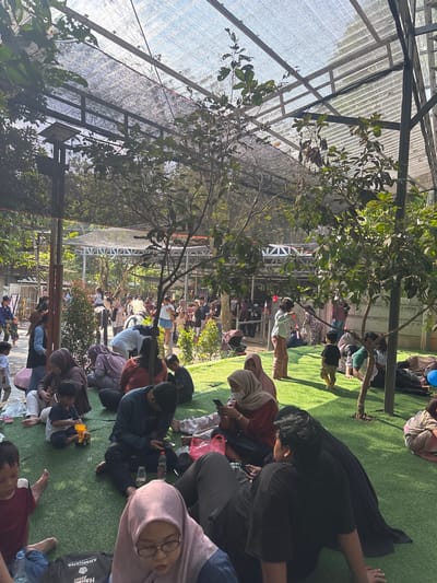 This image depicts a crowded outdoor gathering with numerous people, including adults and children, seated and standing on a carpet of green artificial turf under a shaded canopy. The environment suggests a public space, possibly a park or recreational area, with trees and foliage providing natural shade and contributing to the overall atmosphere. In the background, a white structure with a metal frame is visible, hinting at an enclosed or semi-enclosed area. The people are dressed in casual attire, and many are engaged in various activities such as conversing, using mobile phones, and playing. The presence of a stroller and children playing with toys indicates a family-friendly setting. The lighting suggests it is daytime, with sunlight filtering through the canopy and trees.

The scene is located in Cileduk, Indonesia.