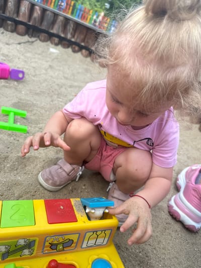 In the city of Donchovci, Bulgaria, a young Caucasian girl with blonde hair tied in a bun is playing in a sandpit. She is wearing a pink t-shirt and pink shorts, with pink shoes. Her hands are covered in sand as she interacts with a colorful yellow toy that has buttons and images of farm animals and bottles. The background shows a wooden fence with colorful vertical posts, likely part of the playground structure. Other toys, including a purple and pink block and a green 'H' shaped toy, are scattered in the sand. The girl is crouched down, intently focused on the toy.