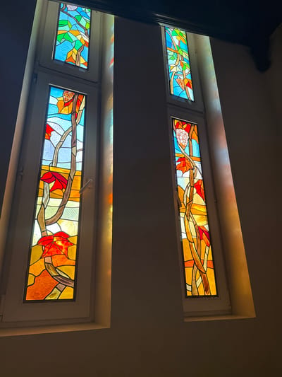 The image shows two tall, narrow stained-glass windows in what appears to be a building. Each window is divided into two vertical sections. The stained glass depicts abstract patterns with organic shapes, predominantly in shades of blue, green, yellow, orange, and red, resembling leaves and branches. Sunlight streams through the windows, casting colorful patterns of light on the adjacent wall. The left window has a white frame and a visible handle on its lower section. The right window also has a white frame and is positioned slightly further away. The walls appear to be painted a neutral, muted color. The overall impression is one of warm, natural light filtering through decorative glass. There is no specific information available to identify this location within the city of Yukki, Russia.