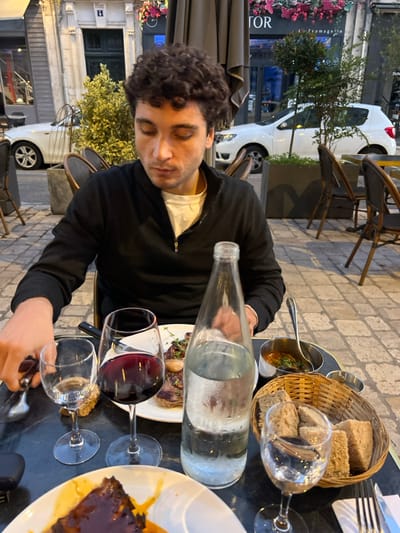 A male individual, appearing to be in his mid-20s to early 30s with curly dark hair, sits at an outdoor table of a restaurant. He wears a black quarter-zip long-sleeved top over a white t-shirt and looks downwards, seemingly focused on the table or his hands, which are near cutlery.
The table, dark in color, is laden with a meal. In front of the man, a white plate holds what appears to be a cooked cut of meat, garnished with herbs and cloves. To his left are two wine glasses, one empty and another containing red wine. A clear glass bottle of water, showing condensation, stands prominently on the table. To his right, a small metal bowl with a spoon holds a dark, saucy dish, and a woven basket contains several slices of bread. A partial view of another plate with a dark, sauced dish is visible in the bottom left.
The scene takes place on a paved street or sidewalk in Orléans, France. In the background, outdoor seating with dark chairs and umbrellas is visible. Behind the seating, two white cars are parked on a street with cobblestone paving. A building facade features a shopfront with a dark frame and glass. An awning above the shop displays the partial text "TOR" and below it, the word "FROMAGERIE" (cheese shop). The building adjacent to the shop has a visible number "4" on its upper floor. Decorative pink and purple flowers hang above the shopfront, adding color to the urban environment.