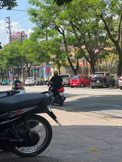 The image depicts a street scene in Lao Cai Ward, Vietnam, on a sunny day. In the foreground, the rear wheel and part of the body of a black motorcycle are visible, parked on a patterned sidewalk. Across the street, several vehicles are parked and in motion. A person wearing a black shirt and a helmet is riding a motorcycle, facing away from the camera. Further down the street, a red car, a black SUV, and a white SUV are parked. On the left side of the street, a person wearing a dark coat and a helmet is riding a motorcycle with large yellow bags attached. Trees with lush green foliage line the street, casting shadows on the pavement. Buildings with colorful facades are visible behind the trees, and utility poles with electrical wires stretch across the sky. The sky is bright blue with scattered white clouds. Yellow leaves are scattered on the sidewalk and the street.