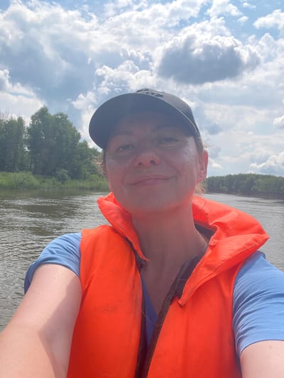 The image is a selfie taken by a woman wearing a blue t-shirt and an orange life vest, with a black baseball cap on her head. She is on a body of water, likely a river, with trees and vegetation lining the banks in the background. The sky is cloudy. The woman is smiling, and her face is partially visible. The scene appears to be outdoors, possibly in a natural or recreational area.