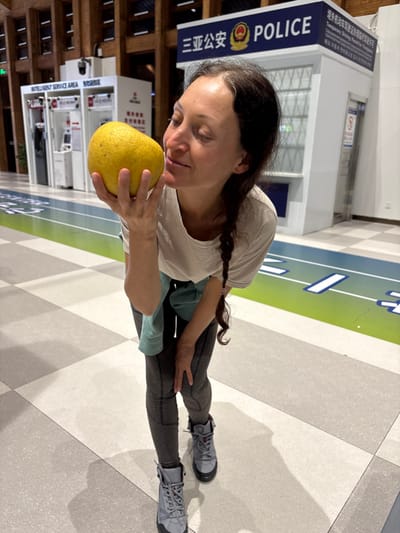An indoor photograph captures a Caucasian female in mid-frame, bent slightly forward, holding a large, spherical yellow fruit to her nose as if smelling it. Her eyes are closed, and she has a slight smile. She wears a light-colored t-shirt, a light blue long-sleeved top tied around her waist, dark grey patterned leggings, and grey hiking-style boots with red laces. The floor consists of light and dark grey checkerboard tiles, traversed by a distinct green and blue striped lane with white markings. In the background, two white modular booths are visible. The booth on the left is labeled "INTELLIGENT SERVICE AREA" in English and Chinese. The booth on the right is a police station kiosk, featuring a prominent dark blue sign with "三亚公安" (Sanya Public Security) and "POLICE" in English, along with the Chinese police emblem. A smaller sign on the right side of the police kiosk mentions "Temporary Driving Permit for Foreigners". The scene is situated within a modern public facility, likely a transit or administrative building, in Tianya District, Sanya, China.