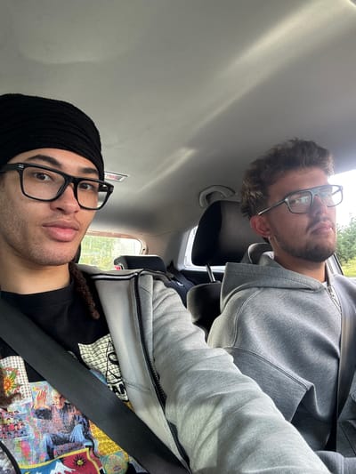 Two men wearing glasses are in the back seat of a car. One man is wearing a black beanie and a t-shirt with a colorful design. The other man is wearing a gray hooded sweatshirt. Both men are wearing seatbelts. The scene is inside the car, no information indicates that the image was taken in Orsay, France.