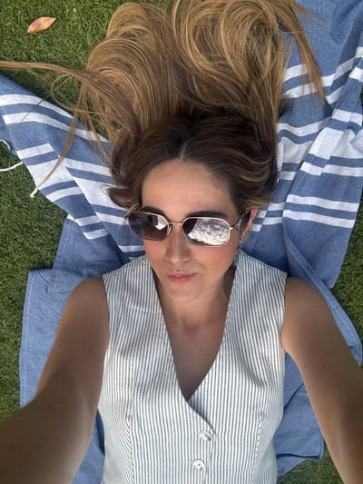 A woman with long brown hair is lying on her back on a blue and white striped beach towel. The towel is spread out on a grassy surface. She is wearing sunglasses, a white sleeveless blouse with blue stripes and a button closure, and a silver necklace. The woman is looking up at the camera. The location is a grassy area in Majadahonda, Spain, at coordinates 40.469 latitude and -3.857 longitude.
