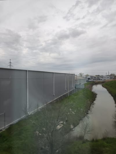 The image depicts an overcast, daytime scene dominated by an industrial or utility landscape in Kanevskaya, Russia. On the left, a long, grey corrugated metal building, likely a warehouse or factory, stretches across the frame. It features several vertical white pipe-like structures along its exterior.
Running parallel to the building on its right, a narrow, murky canal or drainage ditch cuts diagonally across the lower and mid-ground. Its banks are covered with green grass and some sparse, leafless trees or bushes. The canal's water reflects the dull, grey light of the sky.
In the distance, beyond the canal, more grey industrial buildings with dark roofs are visible, along with a few parked cars, suggesting an active industrial or commercial area. A prominent metal electricity transmission pylon stands tall on the left horizon, with power lines extending across the upper portion of the image. The entire sky is covered in light grey, uniform clouds, indicating an overcast day.
The foreground shows significant blurriness due to green foliage, suggesting the photograph was taken from a moving vehicle or through a window. No people or legible text are visible in the image. The overall impression is one of an industrial or semi-rural utility corridor under a dull, cloudy sky.