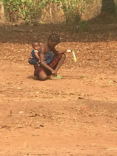 An outdoor scene in Egboje, Nigeria, features an adult female in a squatting posture on reddish-brown dirt ground, with an infant secured to her back by a blue patterned fabric wrap. The adult female, with braided hair, wears a red and black plaid wrap around her lower body, her upper torso bare. She holds a plastic bottle with a white cap and yellow label in her right hand, while her left hand is positioned near a light green object on the ground. The infant, bare-chested and wearing a light-colored undergarment, faces right, peering over the adult's left shoulder. The ground is dry earth with scattered dry vegetation and debris. The background consists of dense green foliage.