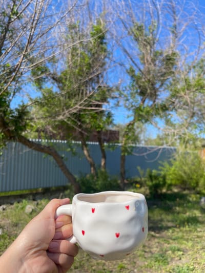 A hand holds a white ceramic mug adorned with small red hearts against an outdoor backdrop. The background consists of tree branches, green foliage, a blue sky, and a section of a corrugated metal fence or wall. The mug is positioned in the right side of the image, obscuring parts of the background. The exact location is unknown, so I cannot say if this is in Verkh-Katunskoye, Russia.