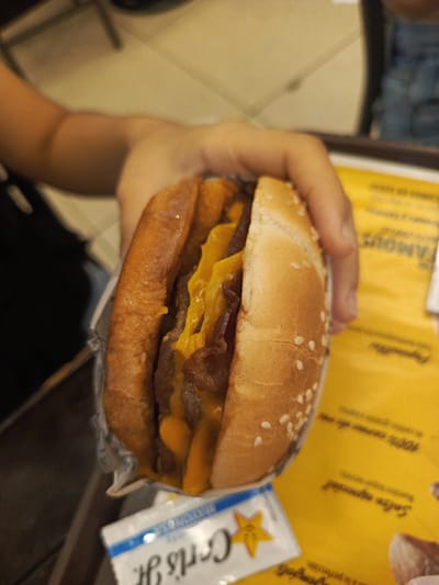 A person is holding a double cheeseburger with bacon in a paper wrapper. The burger is on a yellow tray with writing on it. A small packet of Carl's Jr. mayonnaise is next to the burger. The location is Samborondón, Ecuador, as Carl's Jr. is a franchise restaurant chain with locations in that city.