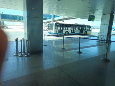 This image captures a scene at Cam Ranh International Airport in Bắc Cam Ranh, Vietnam. A white and blue airport bus, labeled "AGS" and with "NHATRANG" visible above the windshield, is moving along the tarmac. In the background, a jet bridge extends from a terminal building, and mountains are visible on the horizon. The foreground shows a modern airport terminal walkway with polished floors, metal stanchions with ropes, and a sign indicating "G9". The lighting suggests it is daytime with bright sunlight casting reflections on the floor.