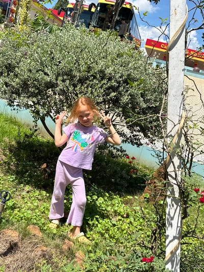 In Ia Hru, Vietnam, a young girl with red hair and a light purple t-shirt and pants poses for a photo outdoors, her hands forming peace signs. She is standing in a grassy area with a large bush behind her. In the background, a yellow and red bus with an open-air upper deck is visible, and a building with a red awning that reads "Ca" can be seen. A concrete pole stands to the right of the frame, with a vine growing around it. The ground is covered in green foliage, and some small red flowers are present in the foreground. The sky is blue with scattered clouds.
