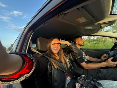 A selfie taken from inside a vehicle shows a woman wearing a black leather jacket, secured with a seatbelt. Her right hand is raised to shield her eyes. A man is visible in the driver's seat. The car's interior includes a sun visor with a warning label. The scene suggests daylight, with a partial view of trees through the window. The location in Babovo, Bulgaria is not evident in this image.