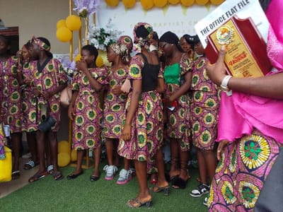 A group of young women, all wearing identical pink and green patterned dresses, stand on green artificial grass in front of a white building. They are posing for a photo. The woman on the far right is holding a plaque which says "THE OFFICE FLAT FILE" and "The Town Baptist Church". She is wearing a pink top with a matching patterned skirt. The location is a church in Yenagoa, Nigeria. The coordinates of the location are 4.931 latitude and 6.267 longitude.