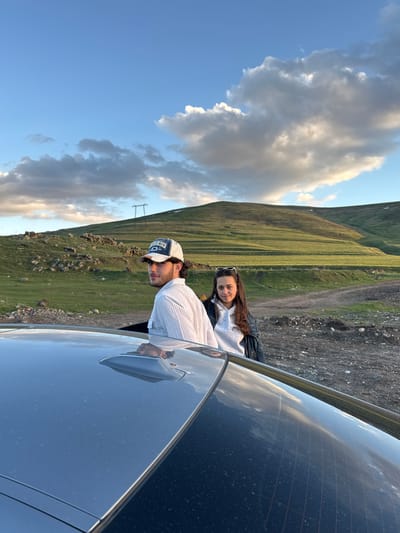 A horizontally oriented medium shot captures two individuals positioned near a dark-colored vehicle, set against a backdrop of mountainous terrain and a sky filled with clouds.
To the left of the frame, a man wearing a horizontally striped light-colored shirt and a baseball cap leans against the car. His face is partially obscured. Adjacent to him, a woman is seen wearing a light-colored sweater underneath a dark jacket. Sunglasses are perched atop her head. The woman is looking toward the camera.
The car occupies a significant portion of the right side of the frame. Behind the figures, a grassy mountain ascends into the horizon, partially obscured by overcast clouds. The scene is set in Hrazdan, Armenia.