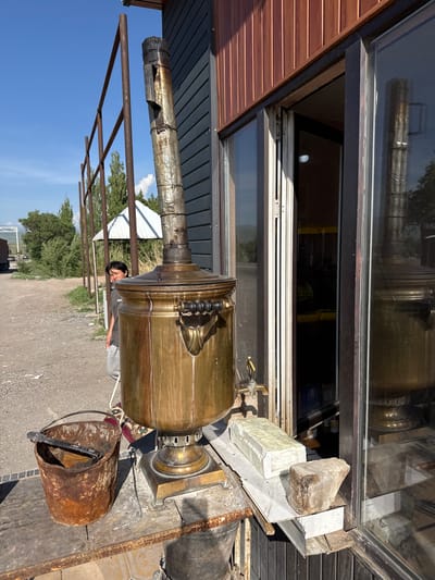 This outdoor scene, captured in Sasyk-Bulak, Kyrgyzstan, features a large, ornate brass samovar as its central subject. The samovar, a traditional tea urn, has a tall, galvanized metal stovepipe extending upwards from its top and a spigot on its side. It rests on a square base on what appears to be an outdoor workstation or platform.

To the right of the samovar, a rusty metal bucket with a handle sits on the ground, containing a dark tool resembling tongs. Behind the samovar, a young person with dark hair, wearing a grey t-shirt and grey pants, is partially visible, looking towards the camera.

The background shows an expanse of dirt ground or a road, with scattered green trees under a clear, bright blue sky with a few wispy clouds. A metal framework, possibly part of an awning or pergola, stretches across the middle ground. To the left, a building with reddish-brown horizontal wooden siding features large glass windows. These windows reflect parts of the outdoor environment and some indistinct shapes inside, but no clear text is visible. The scene is brightly lit by natural sunlight, suggesting a clear day, likely midday given the strong shadows. The overall setting is rustic and appears to be an operational outdoor area, possibly for serving hot beverages.
