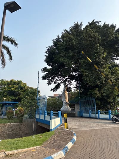 A statue of a robed figure stands near a blue gate and barrier in Bencongan Indah, Indonesia. The statue is white and its face is not visible. The gate is made of blue metal bars and is closed. A yellow and black striped barrier is lowered across the entrance, connected to a rope that hangs from a tall tree. The tree is large and leafy, with dark green foliage. A motorcycle with a rider wearing a helmet is approaching the gate from the right. In the foreground, there is a paved walkway with a blue and white striped curb. A small bridge with blue railings crosses a dry ditch with stone walls. A lamppost stands on the left side of the image, and a palm tree is visible behind it. The sky is clear and blue, and the sun appears to be shining.