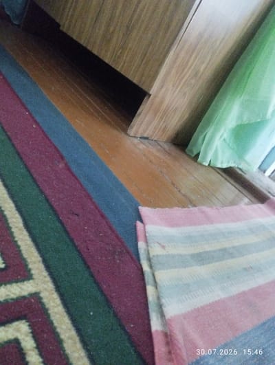 The image captures an interior scene, possibly a domestic dwelling, with a focus on floor coverings and wooden furniture. In the foreground, several striped rugs are visible. One rug, predominantly featuring maroon, dark blue, and green stripes with a cream-colored geometric pattern, lies diagonally from the lower left to the middle of the frame. Another rug, to the right, displays thinner, horizontally oriented stripes in shades of pink, white, light blue, and gray.
The background reveals a wooden floor with a warm, orange-brown hue, showing the grain and natural variations of the wood. A large piece of wooden furniture, likely a cabinet or wardrobe, dominates the upper right portion of the image. Its surface appears to be a lighter wood tone with visible grain patterns, and it is positioned at an angle to the floor. A section of draped light green fabric is visible hanging from or near the furniture, adding a soft element to the otherwise hard surfaces.
The overall lighting suggests an indoor environment, with shadows cast by the furniture. There are no people visible in the image, nor are any specific actions or interactions depicted. The scene is static and presents a snapshot of the room's contents. The timestamp "30.07.2026 15:46" is superimposed in the lower right corner, indicating when the photograph was taken. This image does not provide any specific clues or context to definitively place it in the city of Quyosh, Uzbekistan.