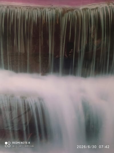 The image is a long exposure photograph of a waterfall, possibly an indoor or artificial one, creating a blurred effect of falling water. In the background, through the water, reflections suggest a person holding a phone and another person wearing a patterned top. The bottom left corner displays the text "REDMI NOTE 8 AI QUAD CAMERA", and the bottom right corner shows the date "2026/6/30" and time "07:42". The scene appears to be in Baýramaly, Turkmenistan, given the device watermark.