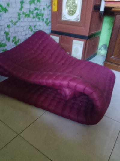 In Merbopuntong, Indonesia, a folded maroon quilted mattress rests on a tiled floor. Behind the mattress, a wall with a green ivy pattern is visible, and to the right, a brown wooden cabinet with decorative elements is present. The lighting is dim, casting shadows and obscuring some details of the environment. There are no individuals or animals present in the scene.