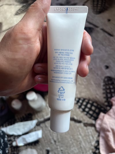 The image shows a close-up of a person's hand holding a white tube of sunscreen. The tube has Korean text and some English text. The background is a blurry mix of a patterned surface and other out-of-focus objects, possibly in a room. The lighting suggests it's daytime, but the specific time of day or weather conditions are not discernible. The focus is on the product and the hand. The visible text indicates "EXP20281223" which is likely an expiration date. The product appears to be a "MOIST FIT SUN CREAM" distributed by Cosmetina Co., Ltd. and manufactured by KOLMAR KOREA CO., LTD. There are also recycling symbols and material indicators visible, "PLASTIC", "LDPE", and "CAP: PP". The scene is intimate and personal, highlighting the product itself.