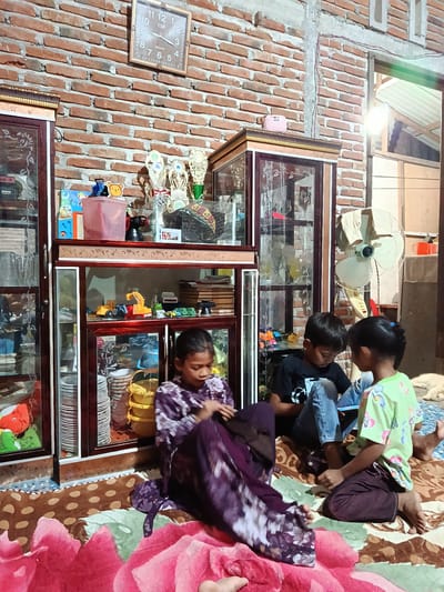 A group of four children are gathered on the floor in a room with a brick wall. Two glass display cabinets filled with various items are prominent features of the room, one on the left and another in the center. The cabinet on the left appears to contain plates and various containers, while the central cabinet displays toys, including toy vehicles, and other decorative items. On top of the central cabinet, there are trophies and what looks like a pink portable speaker. A wall clock hangs on the brick wall, indicating the time.

Three of the children are actively engaged in an activity on the floor. A girl in a purple patterned dress is seated, appearing to be looking at something in her lap. Next to her, two children, a boy in a black t-shirt and jeans, and a girl in a green t-shirt, are looking down at a device, possibly a tablet, held between them. Their focused expressions suggest an engaging activity. Another child's leg is visible in the foreground, suggesting they are also part of the group, though their face is not shown. A fan is visible in the background. The overall atmosphere appears to be one of casual domesticity, possibly in the afternoon, given the lighting. The room seems to be a living space, likely in Indonesia, as indicated by the location context.