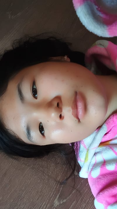 A young girl is lying on her side, looking towards the camera. She has dark hair, dark eyes, and fair skin. She is wearing a pink and white striped robe. The background is a brown wooden surface. The image appears to be taken indoors. There is no indication of the city of Uchkun, Kyrgyzstan in this image.