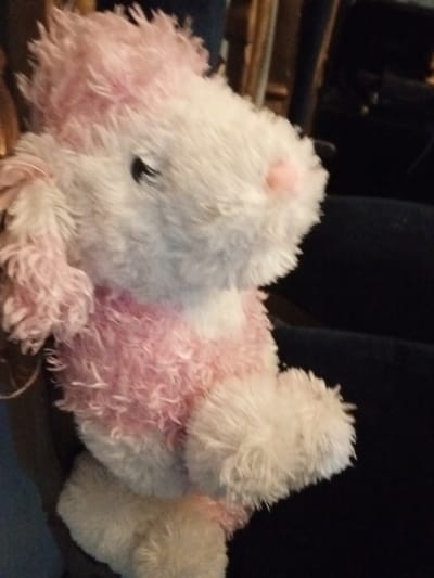 A stuffed animal, likely a bunny, sits upright. It's primarily white with pink accents, including on its head, chest, and one visible paw. The fur is soft and fluffy. Its left ear droops down. A black eye is visible, and its nose is pink. The background is dark, suggesting it's sitting on dark clothing or furniture.