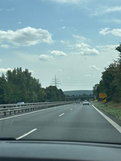 The image shows a view from a vehicle traveling on a multi-lane highway, likely in Germany given the presence of German road signs. The highway has clear lane markings and guardrails. On the left, dense green trees line the road, with a few power lines visible in the distance. On the right, the trees continue, and a yellow road sign with black text is visible, indicating directions and distances to various locations, one of which appears to be "Großwallstadt". Several vehicles are on the highway, including a white convertible, a white van, and other cars. The sky is blue with scattered white clouds, suggesting daytime. The overall scene depicts a typical highway travel environment.