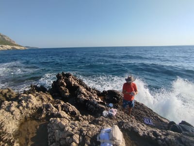A man wearing a cowboy hat, orange t-shirt, and blue patterned shorts stands on rocky terrain near the sea. He is holding something in his hands. Several bags of what appears to be food are on the rocks beside him. A small blue folding chair is also present. Waves crash against the rocks, creating white spray. In the background, the blue sea stretches to the horizon under a clear sky. The rocky landscape suggests a coastal area, likely in the vicinity of Karaburun, Turkey.