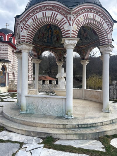 The image depicts an exterior scene in Nepraznentsi, Bulgaria, focusing on a stone rotunda or canopy structure with an integrated fountain. The rotunda features eight white fluted columns with ornate Corinthian-style capitals supporting a series of red and white striped arches. The underside of these arches and the inner dome are adorned with frescoes depicting religious figures and scenes. A multi-tiered white stone fountain, centrally located, presents a cross motif at its apex and lion-head spouts on its upper basin. The rotunda's base is a tiered, circular platform of light-colored stone tiles, carved with floral and geometric patterns, and shows signs of green discoloration, likely algae growth. Surrounding the rotunda are irregularly shaped light gray flagstone pavers separated by patches of sparse grass. In the background, partially visible to the left, is a church constructed of alternating red brick and white stone bands, featuring a red dome capped with black and topped by a golden Orthodox cross. A second building with a red tile roof and stone walls is visible behind the rotunda. The distant background shows a hillside covered with deciduous trees in a leafless state, under an overcast sky. No individuals are present in the scene.