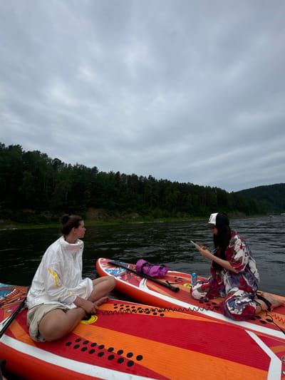 In the city of Mansky, Russia, on a cloudy day, two women are sitting on paddleboards on a wide river. The woman on the left is wearing a white jacket and beige shorts, with her hair pulled back in a bun. She is looking to her right. The woman on the right is wearing a colorful floral shirt and pants and a baseball cap. She is looking down at her phone and holding it in her hands. Both women are sitting with their legs crossed. In the background, there is a dense forest on the riverbank. The water is dark and calm. There are two paddleboards in the foreground, one red and orange with black circles, and another red with orange accents and a blue stripe. A purple bag and a paddle are on the paddleboard with the woman in the floral outfit.