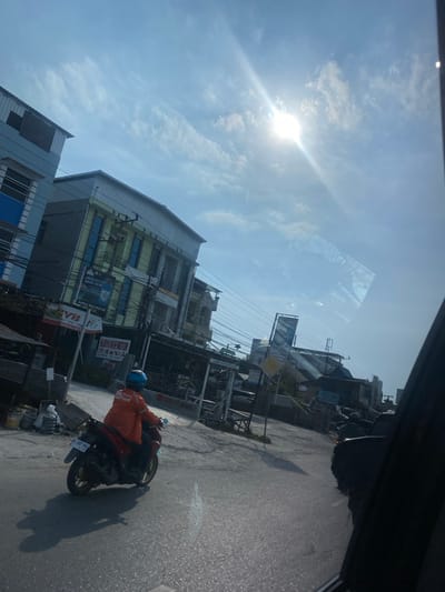 The image depicts a bright, sunny day in Kertak Hanyar I, Indonesia. A person wearing an orange jacket with "Shopee Food" branding and a blue helmet is riding a motorcycle. The motorcycle has a license plate that reads "DA 6107 MAI". The scene shows a street with buildings on the side, including a light blue building and a yellow building with signs. There are power lines strung across the street. The sun is visible in the sky, creating a lens flare effect. The road surface is asphalt, and there is a shadow of the motorcycle and rider cast on the ground. The overall environment appears to be an urban or semi-urban setting.